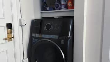Un lavadero pequeño con lavadora de carga frontal, un estante con diversos productos de limpieza y una puerta con cerradura.