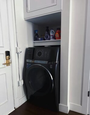 Une petite buanderie équipée d’un lave-linge à chargement frontal, d’une étagère contenant divers produits de nettoyage et d’une porte dotée d’une serrure.