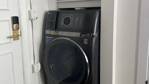 Une petite buanderie équipée d’un lave-linge à chargement frontal, d’une étagère contenant divers produits de nettoyage et d’une porte blanche avec une poignée en laiton.