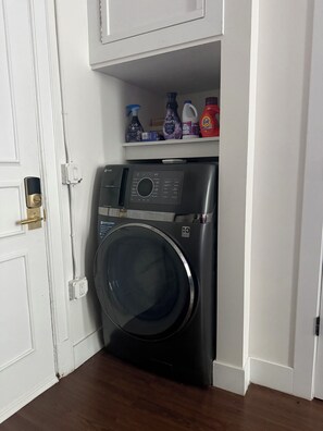Ein kleiner Waschraum mit einer Waschmaschine mit Frontlader, einem Regal mit verschiedenen Reinigungsmitteln und einer weißen Tür mit Messingklinke.