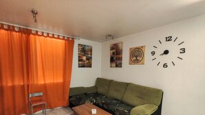 A room with a green sofa, a wooden table, orange curtains, and a wall clock.