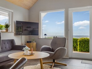 Ein modernes Wohnzimmer mit einem grauen Sofa, einem runden Couchtisch mit Vase und Kerze sowie einem grauen Sessel. Ein Flachbildfernseher ist an der Wand befestigt, und zwei große Fenster bieten Meerblick.