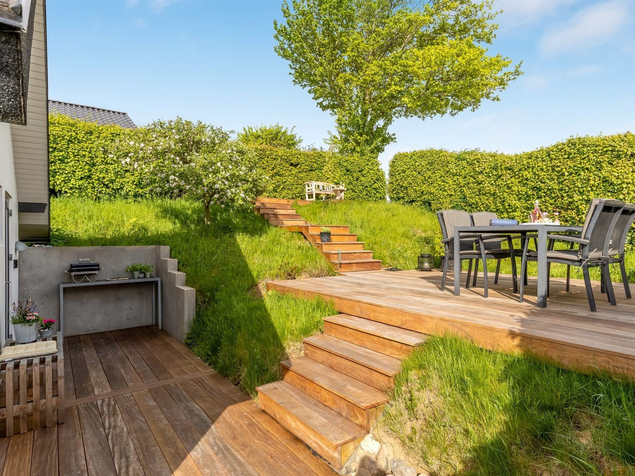 Una terraza de madera con mobiliario exterior, una escalera y un seto al fondo.