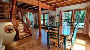 A wooden interior with a staircase, a dining table with chairs, and large windows.