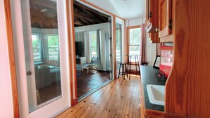 A wooden-floored interior with a kitchen area, a seating area with a table and chairs, and a view through a glass door.