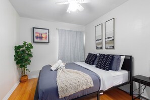 Ein Schlafzimmer mit einem Bett, einem Nachttisch, einem Deckenventilator, einer Topfpflanze und zwei an der Wand befestigten Bildern.
