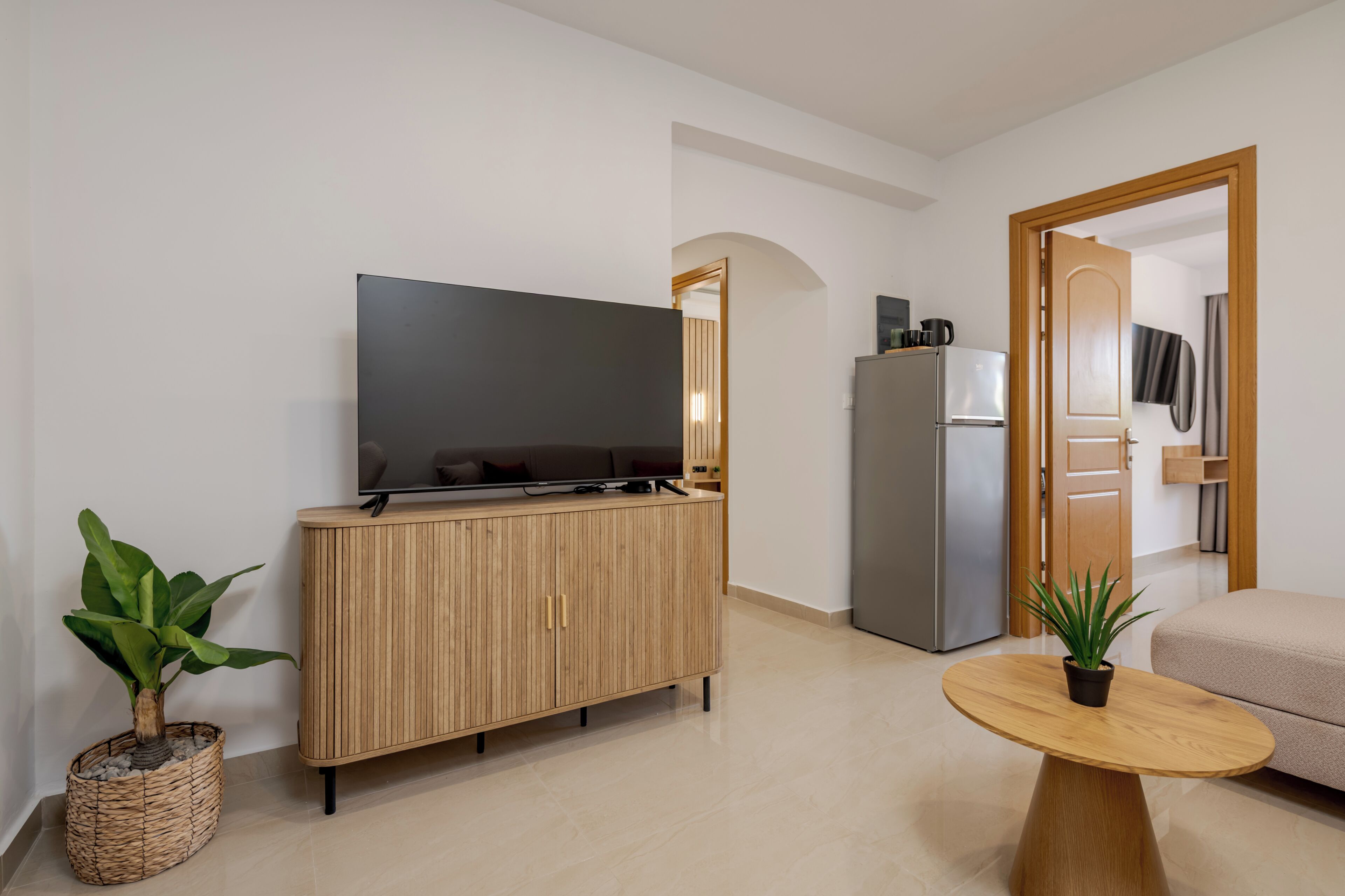 A modern living room with a flat-screen TV on a wooden cabinet, a round wooden coffee table, a beige sofa, and a refrigerator.