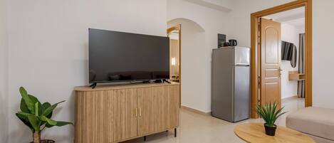 A modern living room with a flat-screen TV on a wooden cabinet, a round wooden coffee table, a beige sofa, and a refrigerator.