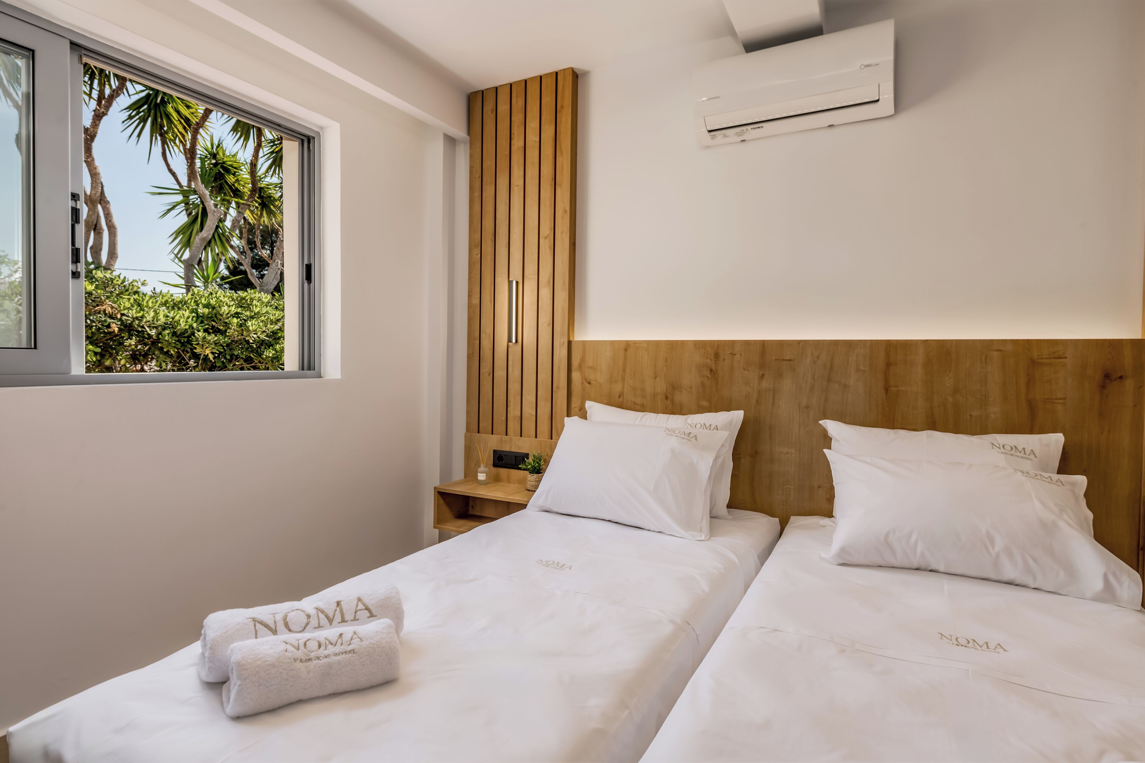 Ein Hotelzimmer mit zwei Betten, einem Holzkopfstück, einer Klimaanlage und einem Fenster mit Blick auf Palmen.
