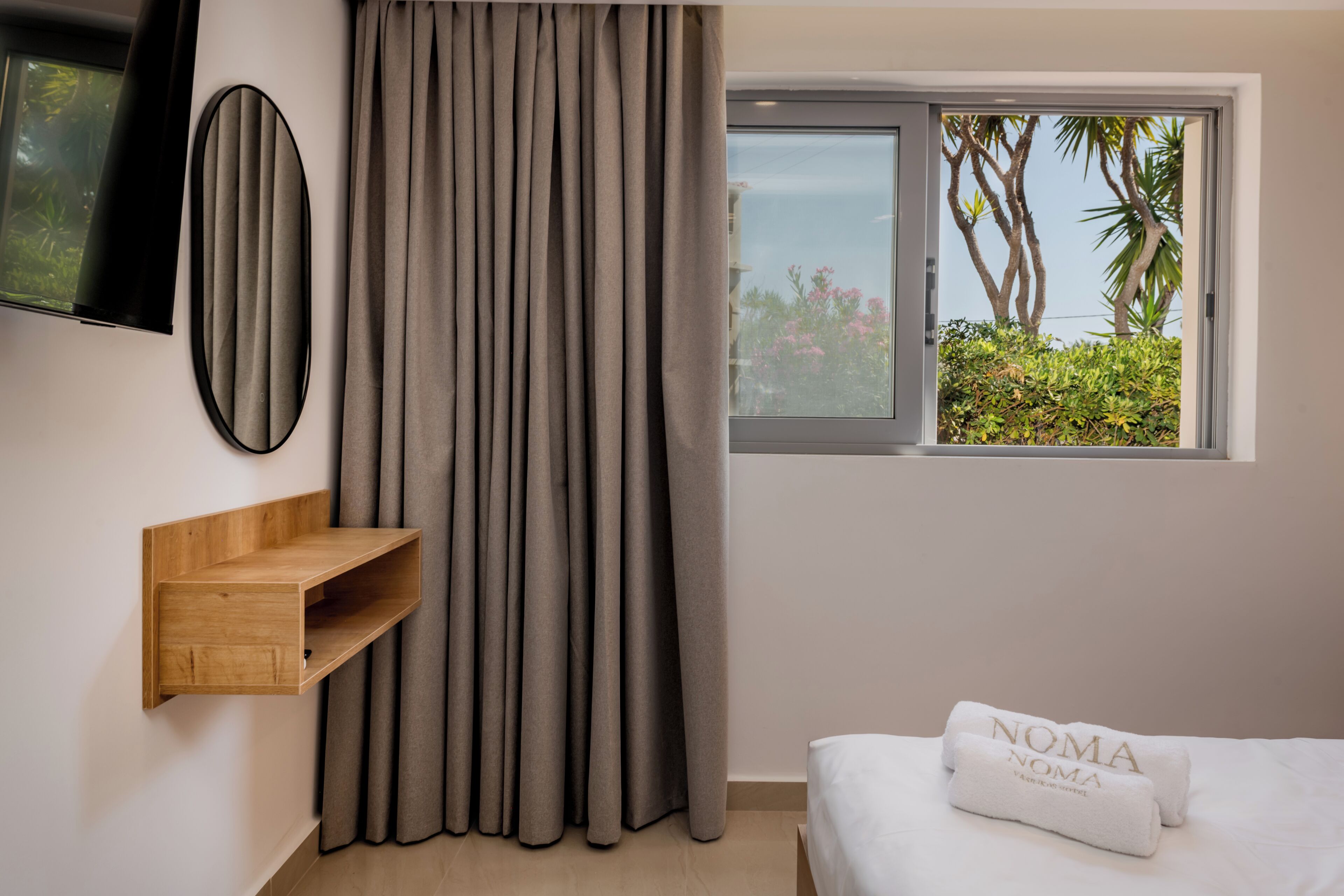 A bedroom with a bed, a TV mounted on the wall, a window with a view of trees, and a wooden shelf.