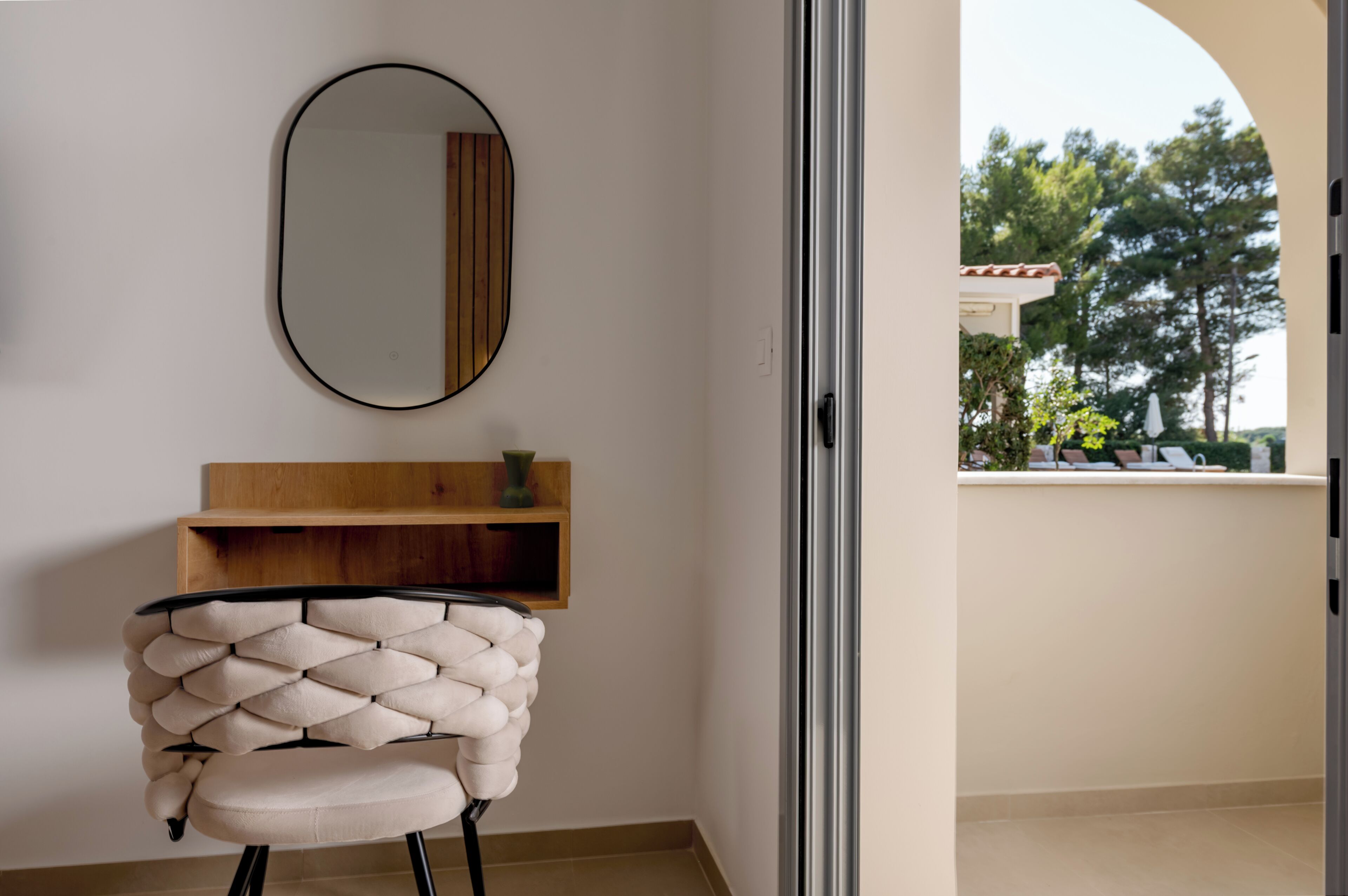 A room with a chair, a mirror, a wooden shelf, and a view of a balcony with outdoor furniture.