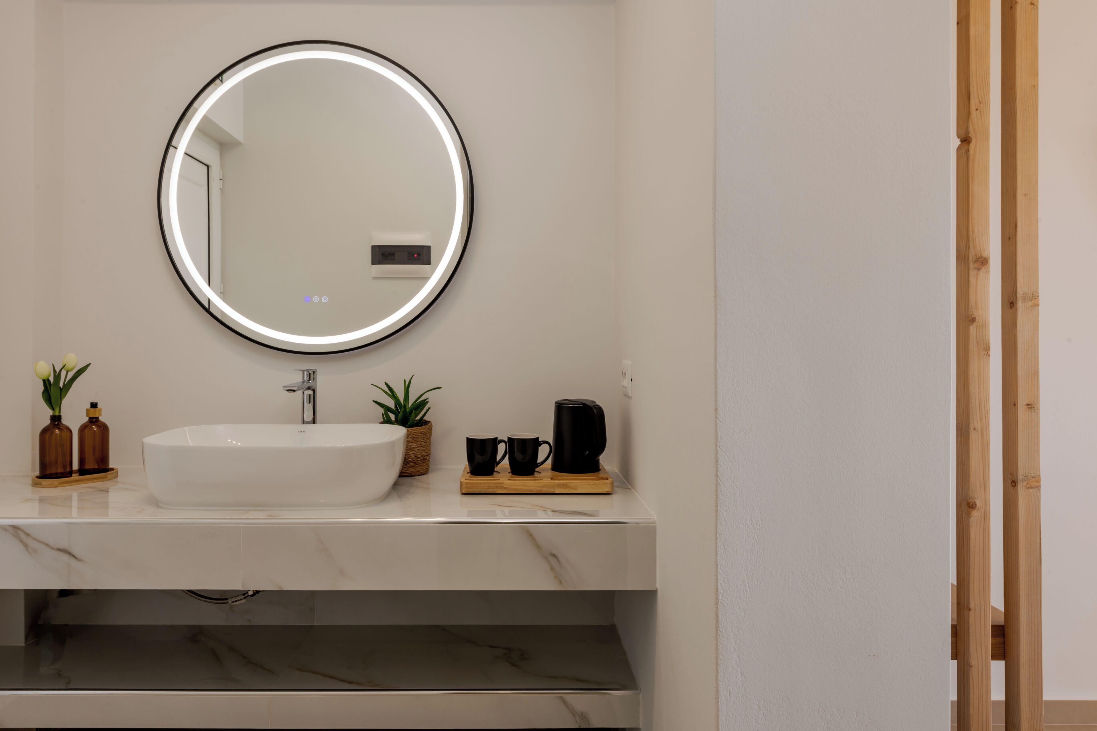 Un baño con espejo redondo, lavamanos blanco y estante de madera con dos botellas pequeñas y una planta.