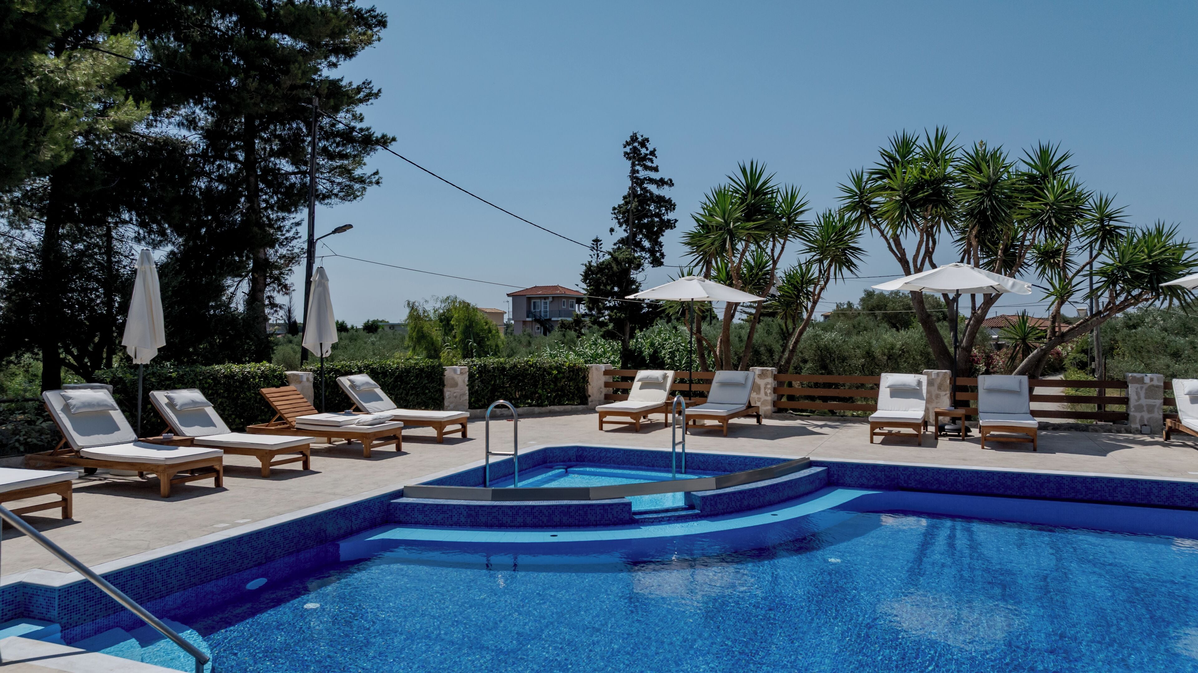 A pool area with lounge chairs and umbrellas, surrounded by trees and a clear sky.