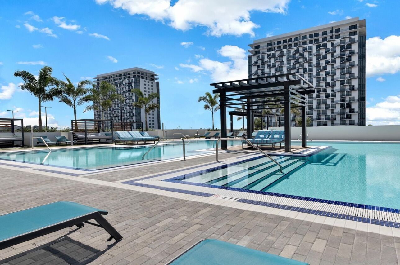 En swimmingpool med solstolar och en pergola, omgiven av palmer och moderna byggnader.