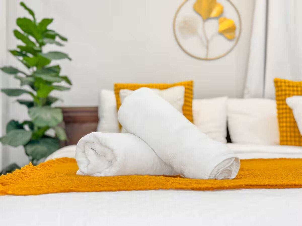 A neatly made bed with white linens and a yellow throw blanket, a plant to the left, and a wall with a circular decoration.