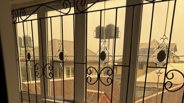 Visão através de grades metálicas ornamentadas, com piso de azulejos e um vislumbre de edifícios ao fundo.