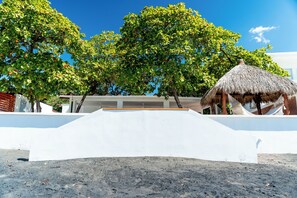 A beachside view with a white building, a thatched-roof structure, and lush green trees.