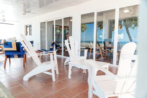 A sunlit room with white chairs and a table, large windows, and a view of the sea.