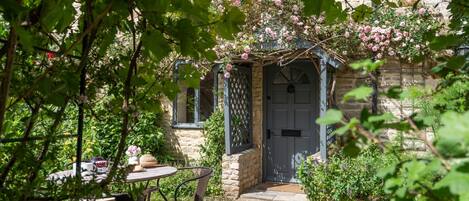 Un bâtiment en pierre avec une porte, une table entourée de chaises et un jardin fleuri.