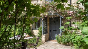 A stone building with a door, a table with chairs, and a garden with flowers. - Rose Cottage Oddington - Countryside  Retreat, 2 bedroom, Lower Oddington (Oddington)
