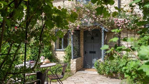 A stone building with a door, a table with chairs, and a garden with flowers. - Rose Cottage Oddington - Countryside  Retreat, 2 bedroom, Lower Oddington (Oddington)