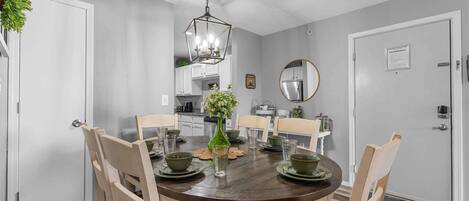 A dining room with a round table, chairs, a chandelier, and a kitchen in the background.