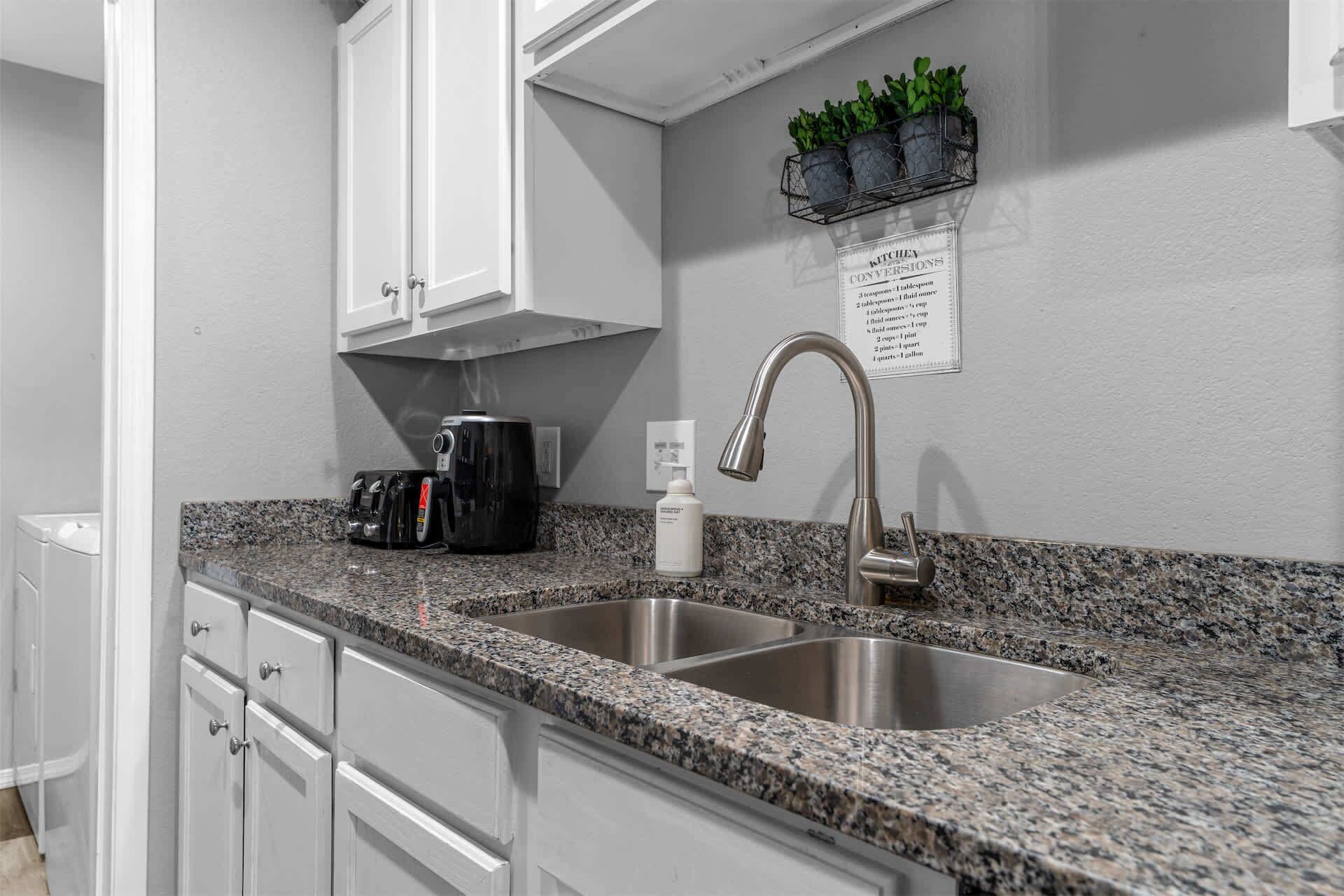 Una cocina con encimeras de granito, lavamanos de acero inoxidable, armarios blancos, una cafetera, una botella de detergente y un cartel con conversiones de medidas culinarias.