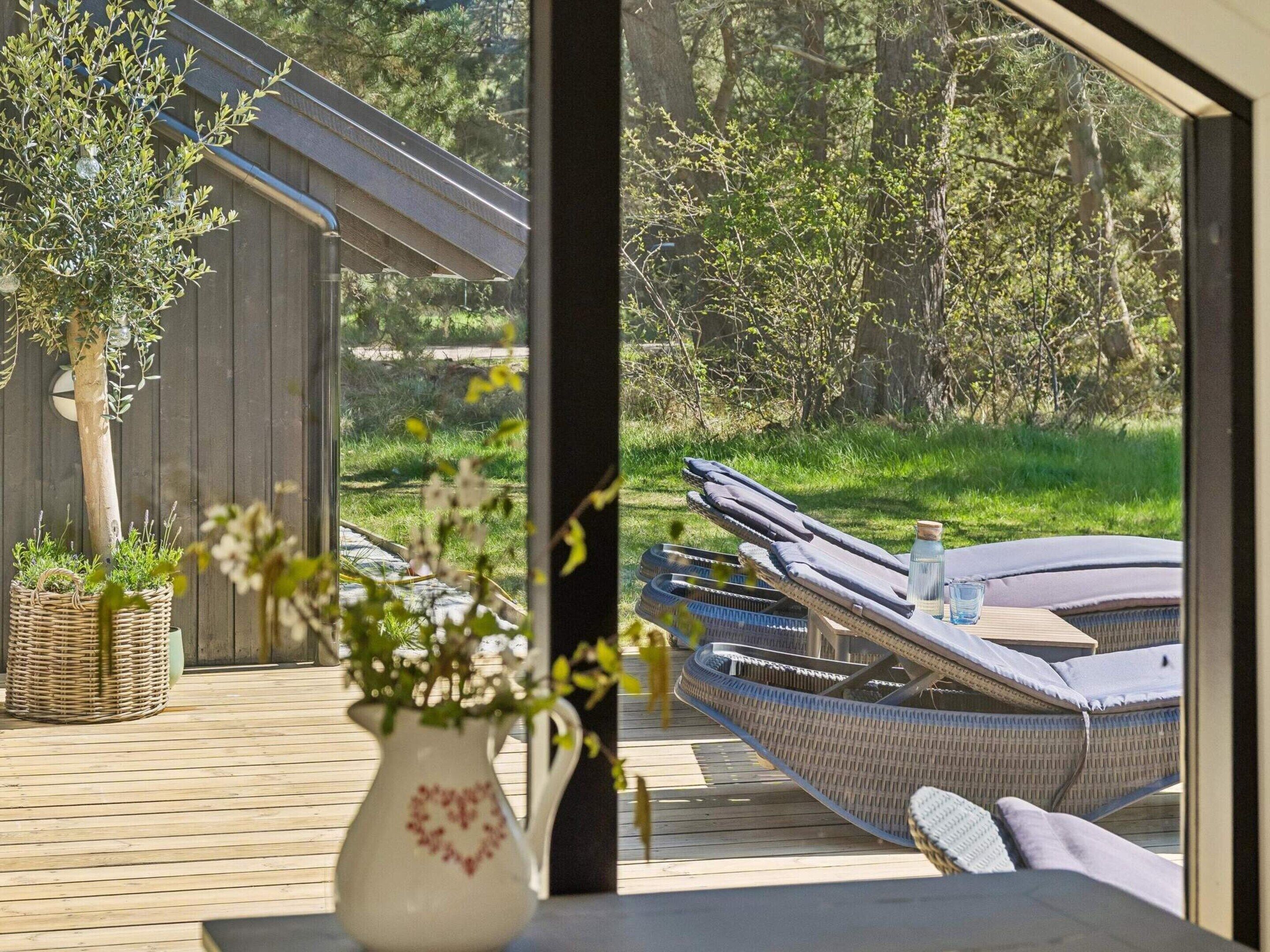 Une terrasse en bois, dotée de chaises longues et d’une table en osier, offre une vue sur un jardin arboré et un bâtiment.