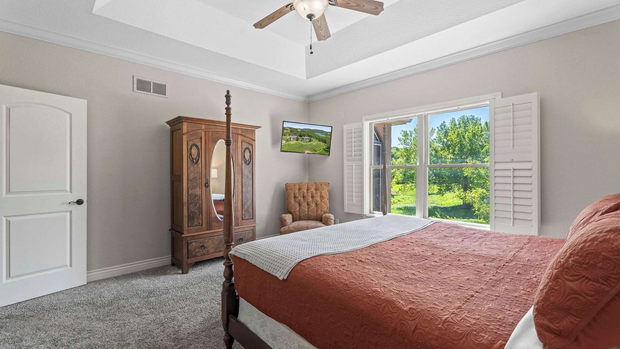 A bedroom with a bed, a wooden wardrobe, a chair, a ceiling fan, and a window with a view of greenery.