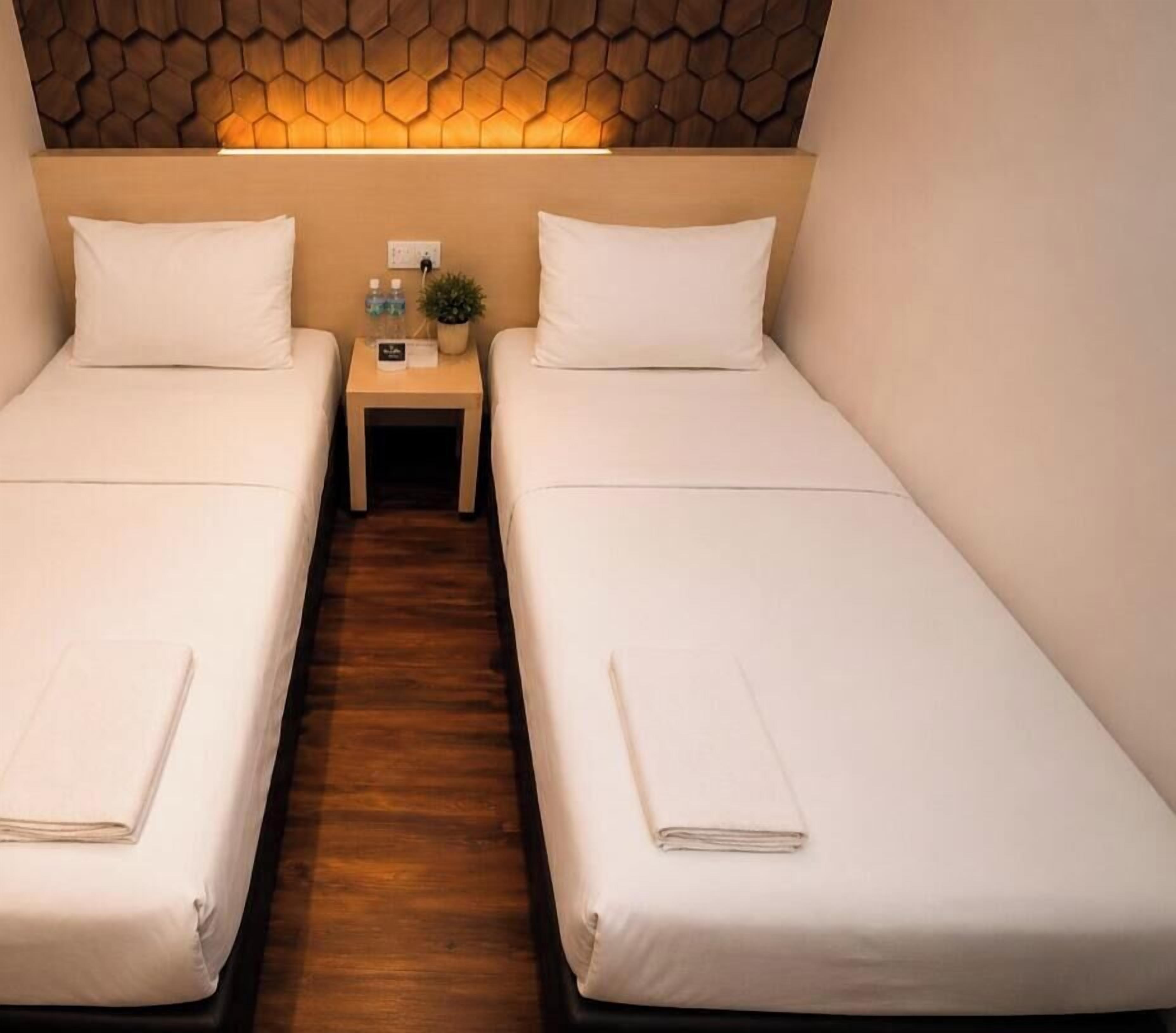 Quarto duplo com duas camas, piso de madeira, luminária na parede e uma pequena mesa com uma planta e uma garrafa de água.