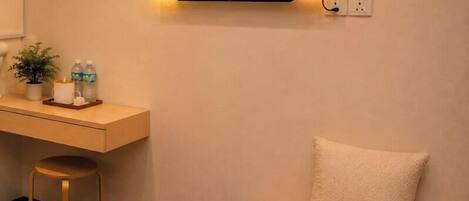 Habitación de hotel con una cama, un escritorio con silla, un televisor montado en la pared, un almohadón y una toalla enrollada sobre un banco.