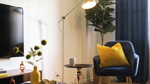 A modern living room with a flat-screen TV, a black armchair with yellow pillows, a small round table, a floor lamp, and a potted plant.