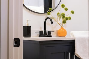 Ein Badezimmer mit einem schwarzen Wasserhahn, einem weißen Waschbecken, einem Spiegel und einer Vase mit grünen Blumen.