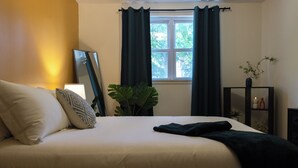 A neatly made bed with pillows, a bedside plant, a window with curtains, and a small shelf with items.
