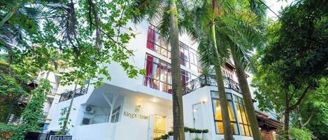 Un hotel con fachada blanca, varios balcones y abundante vegetación a su alrededor.