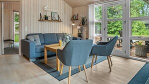 Ein modernes Interieur mit Holzboden, einer Couch, zwei Sesseln, einem Tisch und einem großen Fenster mit Blick auf Grünflächen.