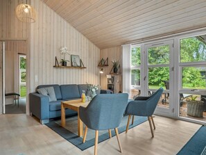 Un interior moderno con techo de madera, un sofá, dos sillas, una mesa y un amplio ventanal con vistas a la vegetación.
