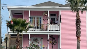 A two-story pink house with a balcony, a front porch, and palm trees. - Shelby by the Seashore C1 (Galveston)