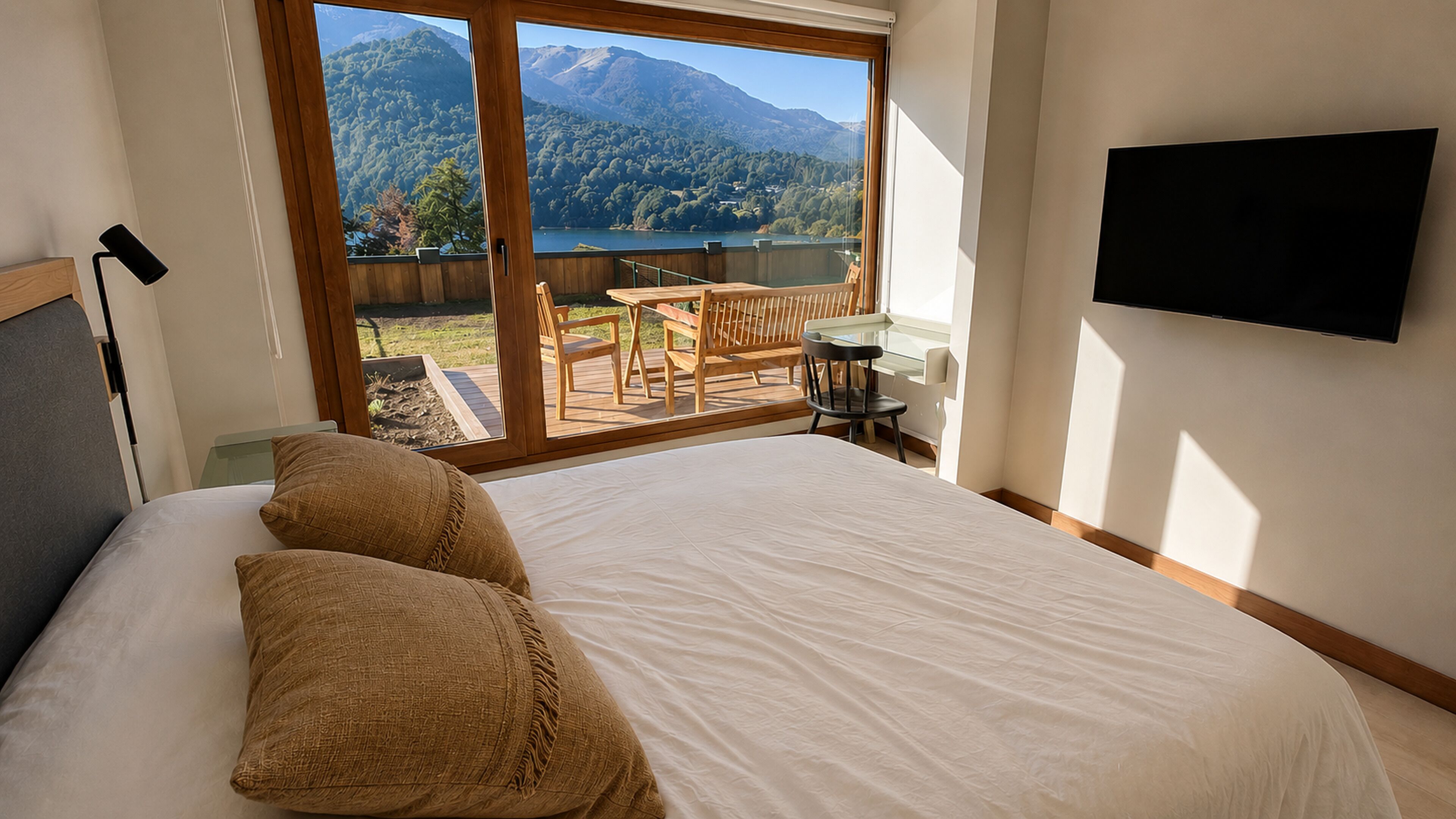 A bedroom with a large bed, a TV mounted on the wall, and a balcony with a view of mountains and a lake.