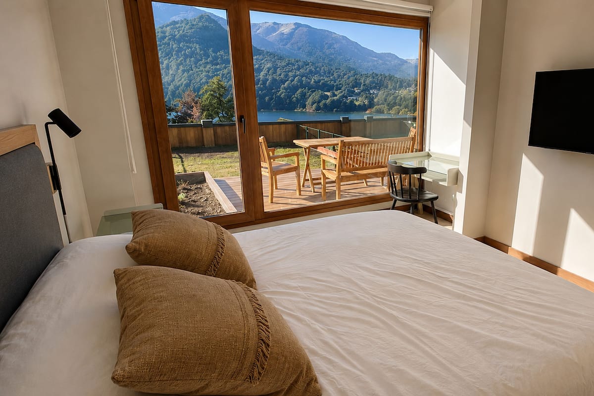 Un dormitorio con una cama grande, un televisor montado en la pared y un balcón con vistas a montañas y un lago.