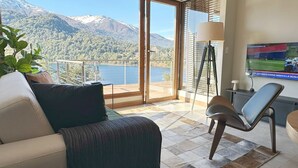 A modern living room with a sofa, a chair, a TV, and a view of a lake and mountains.