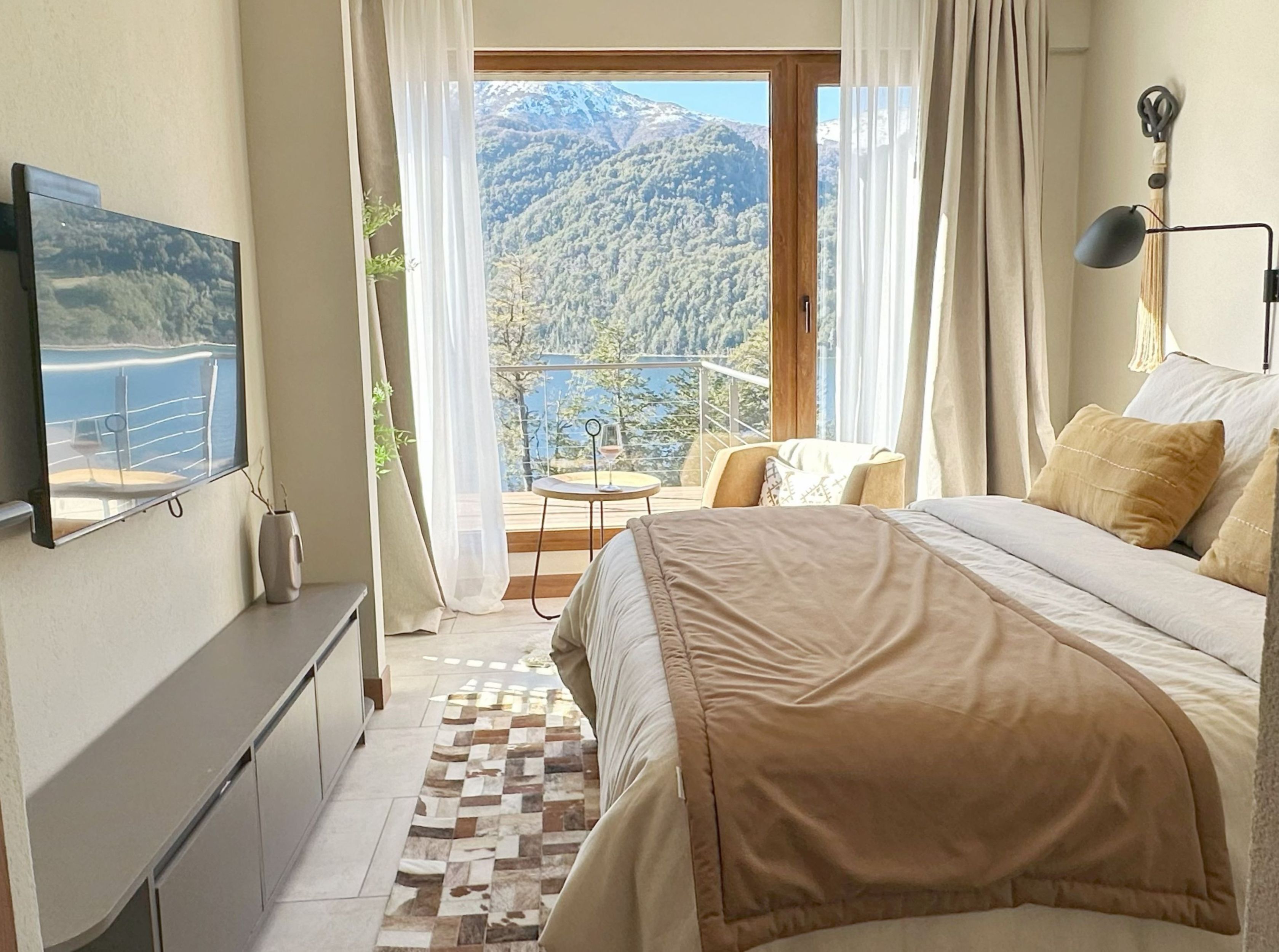 A bedroom with a bed, a TV, a balcony with a table and chairs, and a view of mountains.