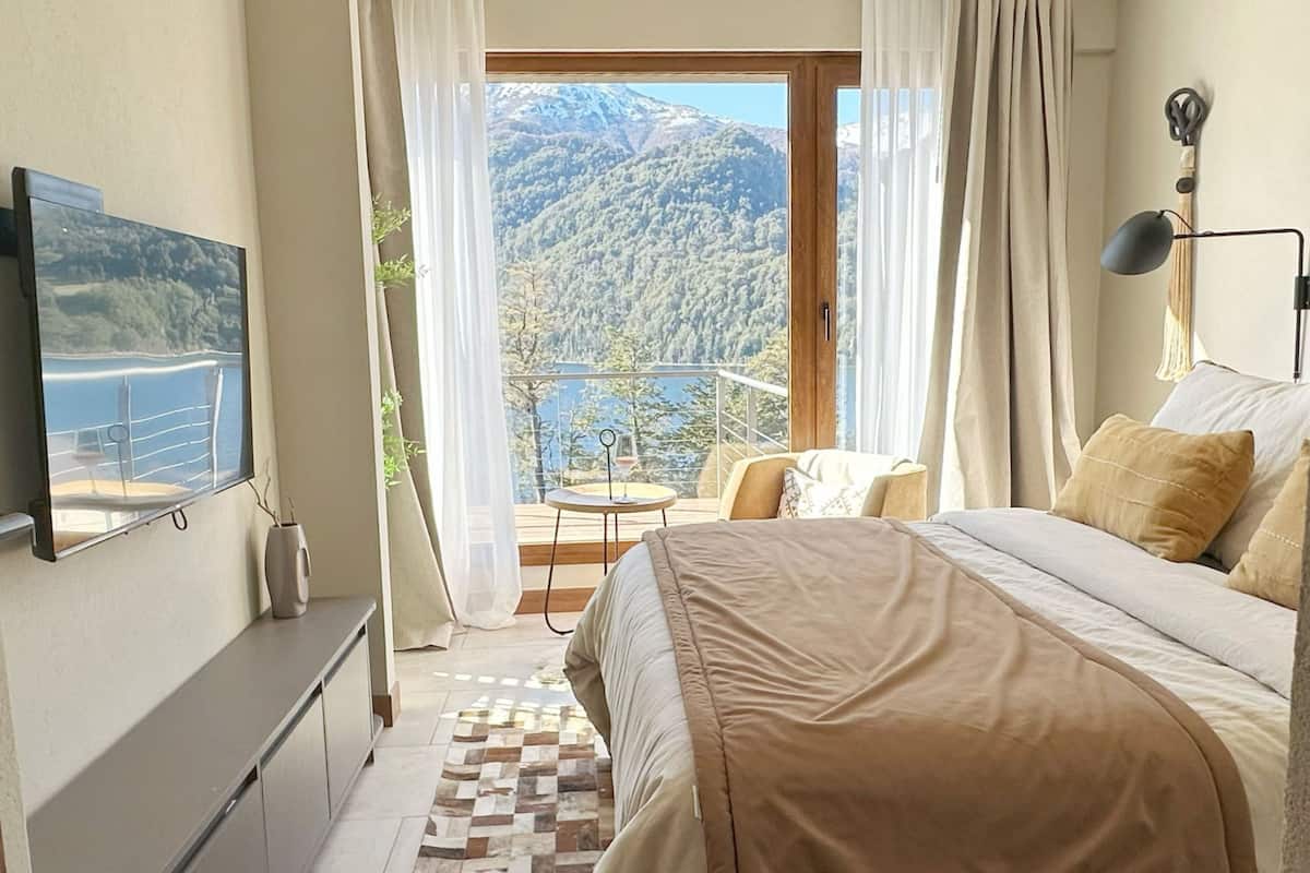 Un dormitorio con cama, televisión, balcón con mesa y sillas, y vistas a la montaña.