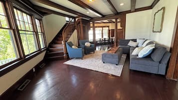 A spacious living room with a staircase, wooden floors, and large windows.