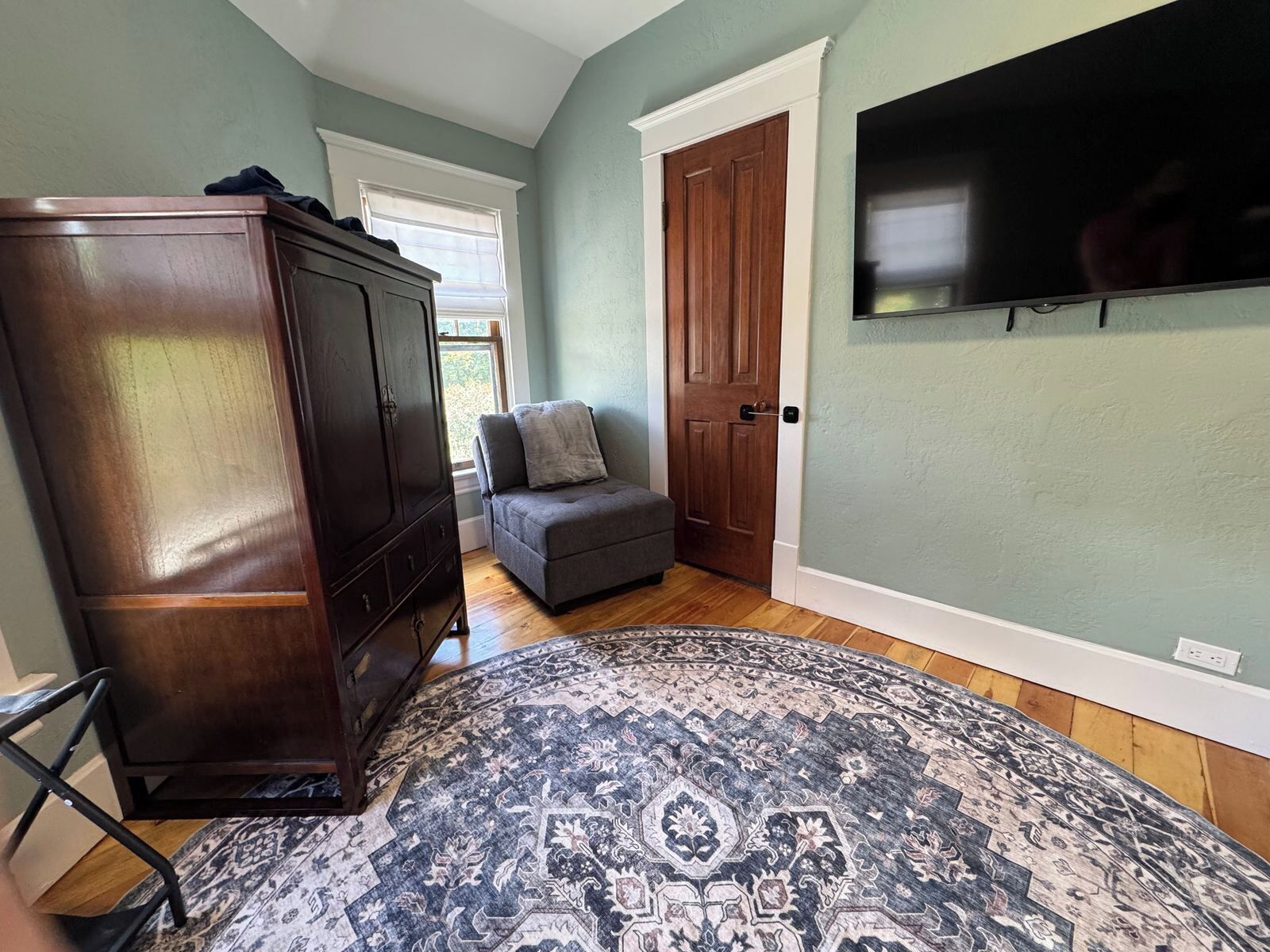 A room with a wooden wardrobe, a grey sofa, a patterned rug, and a television mounted on the wall.