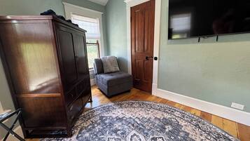 A room with a wooden wardrobe, a grey sofa, a patterned rug, and a television mounted on the wall.
