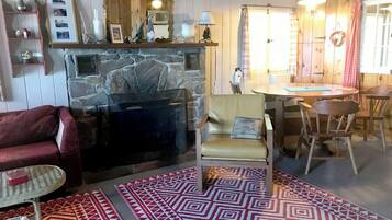 Un salon chaleureux avec une cheminée en pierre, un canapé rouge, un tapis à motifs et un coin repas avec des chaises et une table en bois.