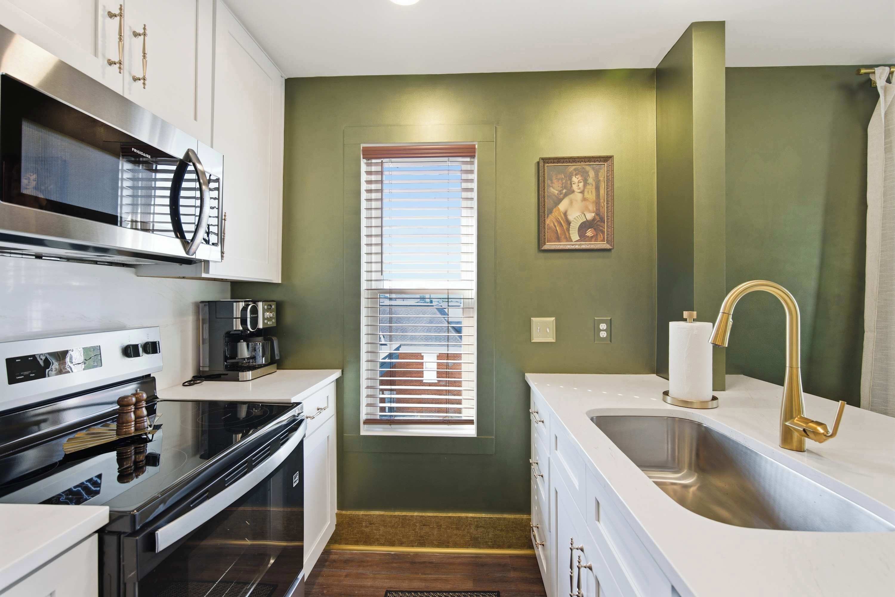A kitchen with a built-in oven, a coffee maker, a sink with a gold faucet, and a window with blinds.