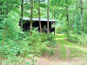 Una cabaña rodeada de un bosque denso, con una escalera visible que lleva a la entrada.