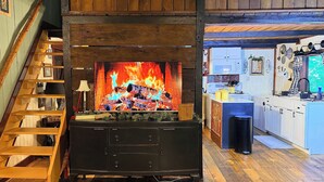 A cozy living room with a fireplace, wooden staircase, and a kitchen area.
